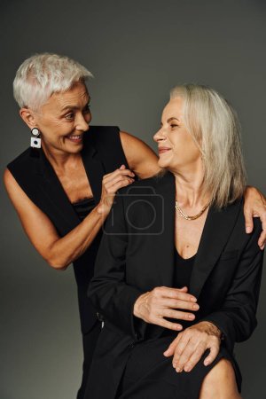 joyful mature ladies in black fashionable attire smiling at each other on grey, lifelong friendship