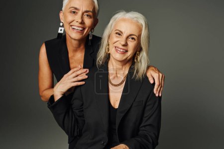 fröhliche und elegante ältere Damen in schwarzer Freizeitkleidung blicken vor grauem Hintergrund in die Kamera