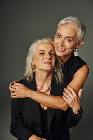 Foto de Señora madura de moda en ropa negra abrazando elegante amiga femenina en gris, envejecimiento feliz - Imagen libre de derechos