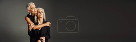 fashionable and dreamy senior lady hugging female friend and looking away on grey backdrop, banner