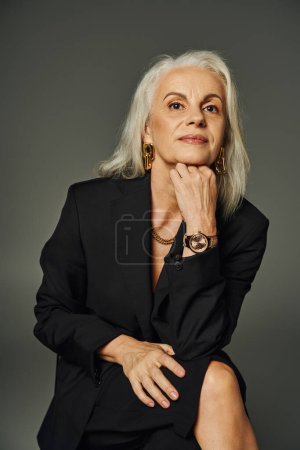 Foto de Señora mayor pensativa en traje negro de moda sentado y mirando hacia otro lado en gris, envejeciendo con gracia - Imagen libre de derechos