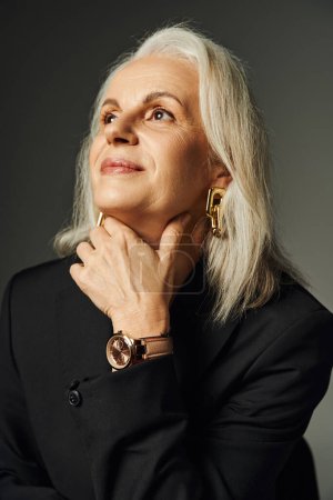 portrait de dame âgée rêveuse et élégante en montre-bracelet et accessoires dorés regardant loin sur gris