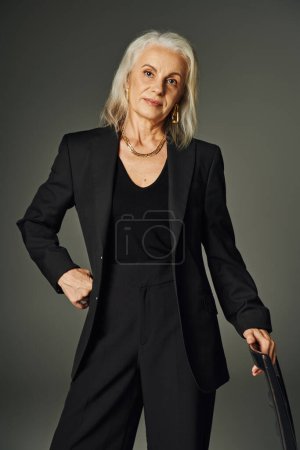 elegant senior model in black clothes and golden accessories near chair with hand on hip on grey