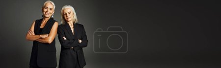 happy senior fashionistas in black classic attire posing with folded arms on grey backdrop, banner