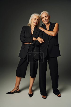 Foto de Longitud completa de alegres modelos senior en negro elegante ropa de pie y sonriendo a la cámara en gris - Imagen libre de derechos