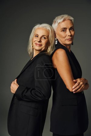 senior models in black classic clothes standing back to back and looking at camera on grey