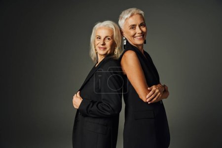 heureux modèles féminins seniors en costume classique noir posant dos à dos et souriant à la caméra sur gris