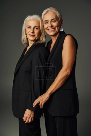 Photo for Vanity fair style, joyful senior women in black classic attire looking at camera and posing on grey - Royalty Free Image