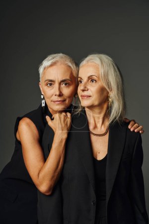 elegante Seniorin in schwarzem Gewand umarmt Schultern einer Freundin und blickt in die Kamera auf grau