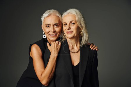 cheerful senior lady in black classic attire hugging shoulders of female friend on grey, happiness