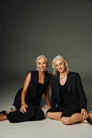 Téléchargez les photos : Dames matures à la mode en vêtements noirs assis et souriant à la caméra sur gris, mode classique - en image libre de droit