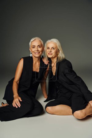 modelos senior sonrientes en traje elegante negro sentado sobre fondo gris, envejecimiento elegante y amistad