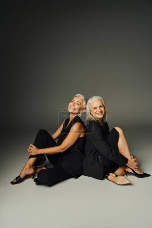 cheerful senior female friends in black attire sitting back to back on grey, fashionable aging