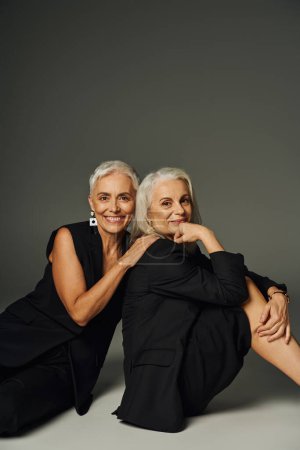 modische Seniorinnen in schwarzen Kleidern sitzen und lächeln in die Kamera auf grau, Positivität