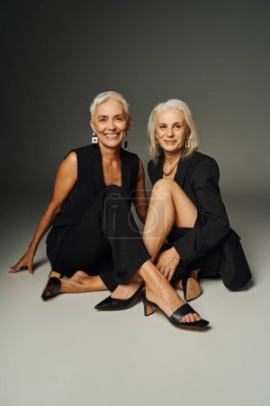 Positif senior trendsetters en tenue noire assis et souriant à la caméra sur gris, mode classique