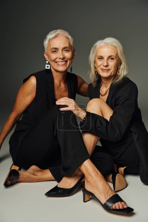 Charmante elegante Seniorinnen in schwarzen Kleidern sitzen und lächeln in die Kamera auf grauem, stylischem Altern