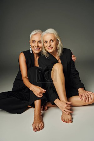 fröhliche und trendige barfüßige Seniorinnen in schwarzer Kleidung, die in die Kamera schauen, während sie auf grau sitzen