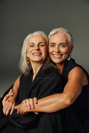 happy aging, cheerful and stylish female friends in black attire embracing and posing on grey