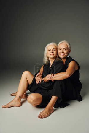 Barfüßige Seniorinnen in schwarzen Klamotten sitzen und blicken in die Kamera auf grau, altersgerecht