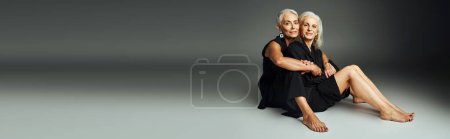 Photo for Fashionable barefoot senior models in black casual attire sitting on grey backdrop, banner - Royalty Free Image