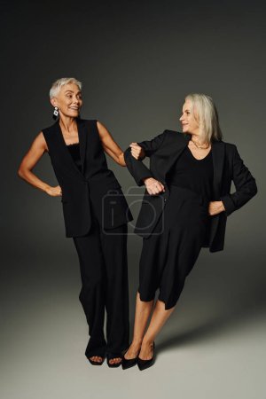 senior female friends in black trendy wear having fun and smiling at each other on grey, full length