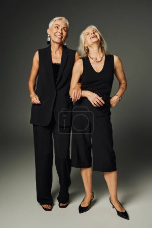 full length of excited mature ladies in black classic attire standing and laughing on grey backdrop