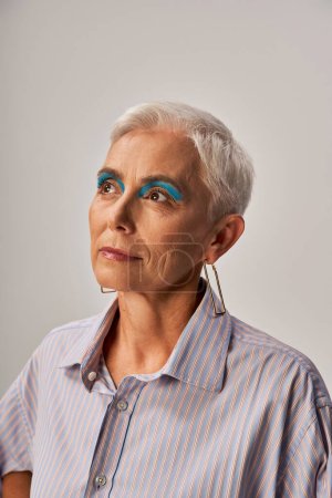 élégant modèle senior rêveur avec cheveux courts argentés et eye-liner bleu regardant loin sur gris, portrait