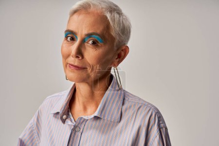 Téléchargez les photos : Portrait d'élégante dame mature aux cheveux courts argentés et au maquillage audacieux souriant à la caméra sur gris - en image libre de droit
