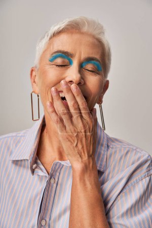 mujer madura excitada con maquillaje audaz riendo con los ojos cerrados y cubriendo la boca con la mano en gris