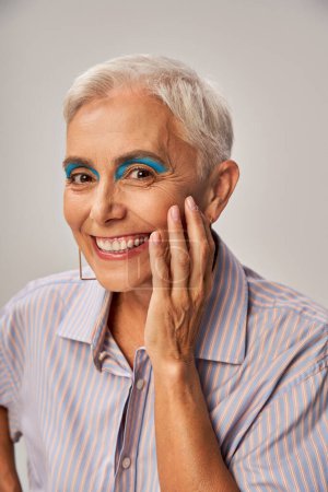 portrait de mannequin senior élégant aux cheveux argentés courts et au maquillage audacieux souriant à la caméra sur gris