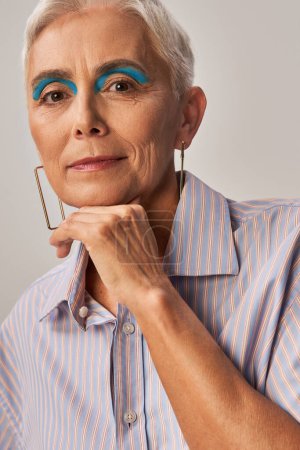 trendige reife Dame mit kurzen silbernen Haaren und blauem Eyeliner posiert mit händennahem Kinn auf grau