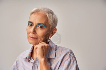 modelo senior de moda con maquillaje audaz y pelo corto plateado mirando a la cámara en gris, pancarta