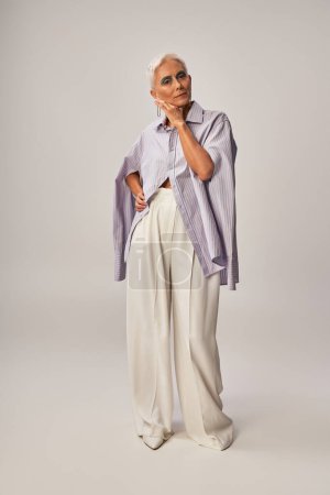vieillissement à la mode, pleine longueur de la femme âgée en chemise à rayures bleues et pantalon blanc debout sur gris