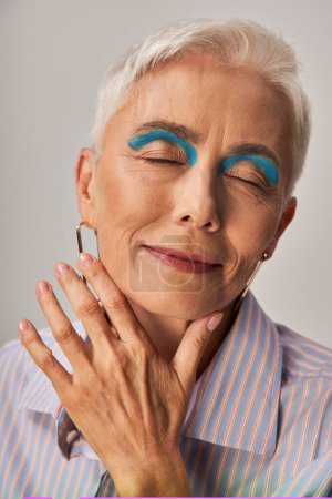 femme mature positive avec les cheveux courts argentés et eye-liner bleu posant avec les yeux fermés sur gris