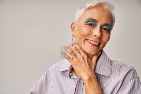 fröhliche ältere Dame mit blauem Eyeliner und kurzen silbernen Haaren blickt vor grauem Hintergrund in die Kamera