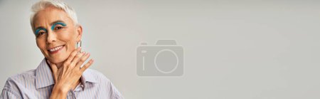 Photo for Joyful stylish mature model with blue eyeliner and short silver posing with hand near chin on grey - Royalty Free Image