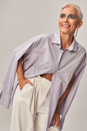 joyeuse femme âgée en chemise à rayures bleues tenant la main dans la poche et regardant loin sur gris