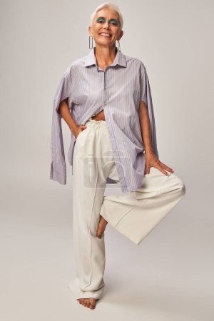 joyful and barefoot mature lady in blue striped shirt posing on one leg with hand in pocket on grey