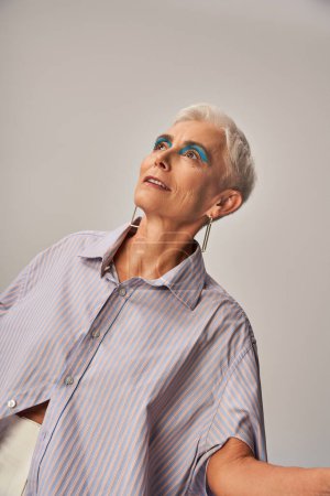 dreamy and fashionable senior woman with bold makeup and short silver hair looking away on grey