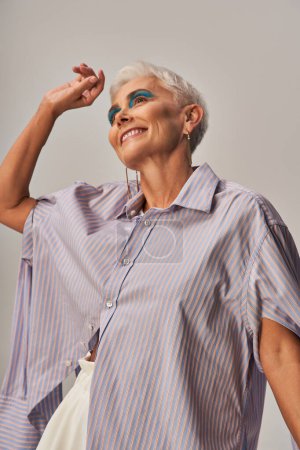 Foto de Mujer mayor alegre y de moda con maquillaje audaz y pelo corto plateado mirando hacia otro lado en el fondo gris - Imagen libre de derechos