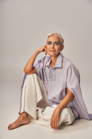 fashionable barefoot senior woman in blue striped shirt and white pants sitting on grey backdrop