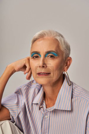 Trendy Senior Model mit kurzen silbernen Haaren und blauem Eyeliner blickt in die Kamera auf grau, Portrait