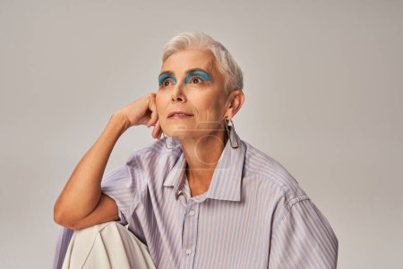 pensive and stylish senior woman in blue striped shirt and bold makeup looking away on grey