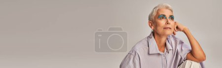 Téléchargez les photos : Portrait de femme mature rêveuse en chemise à rayures bleues et maquillage audacieux regardant loin sur gris, bannière - en image libre de droit