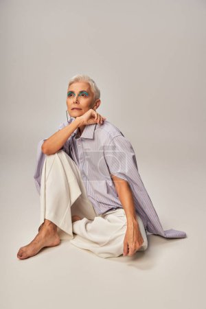 barefoot and dreamy senior model in blue striped shirt and pants sitting and looking away on grey