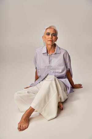 Barfüßige und selbstbewusste Seniorin in trendiger Freizeitkleidung blickt in die Kamera, während sie auf grau sitzt