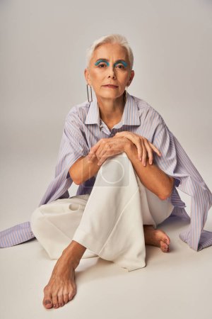 barefoot and fashionable senior lady in stylish casual wear sitting and looking at camera on grey