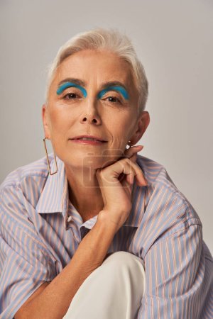 mature lady with bold makeup posing in blue striped shirt and smiling at camera on grey