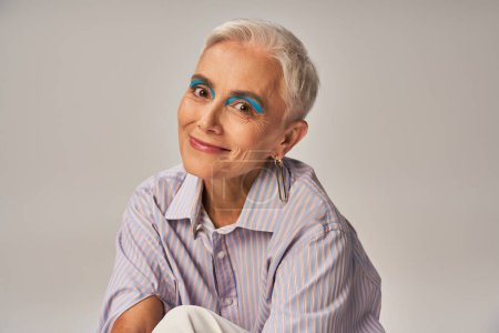 positive mature lady with bold makeup in blue striped shirt smiling at camera on grey, banner