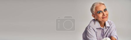 Photo for Fashionable senior lady with bold makeup in blue striped shirt smiling at camera on grey, banner - Royalty Free Image